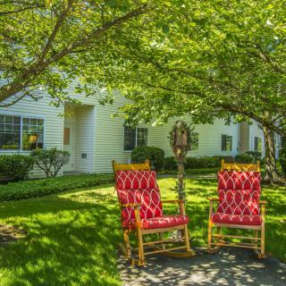A peaceful garden setting with two red rocking chairs under a tree, in front of a white house.