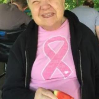 A smiling person wearing a pink ribbon t-shirt and black jacket, holding a slice of watermelon.