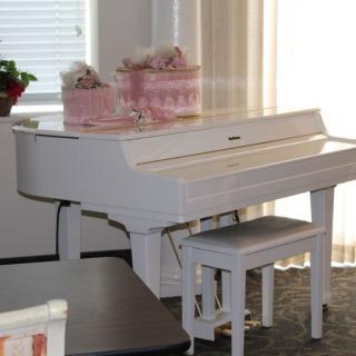 A white piano with a matching bench, adorned with pink floral decorations, situated in a sunlit room.