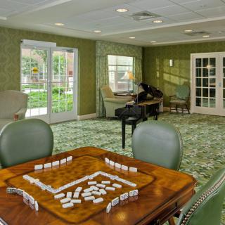 A cozy living room with green walls, featuring a table set for a game of dominoes, surrounded by comfortable chairs and large windows.