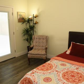 A cozy bedroom with a decorative bedspread, an armchair, a floor lamp, and a potted plant next to a glass door.