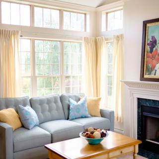 A cozy living room with a light blue sofa, accented by yellow and blue cushions. Large windows with sheer curtains allow natural light to fill the space. A wooden coffee table sits in front of the sofa, topped with a decorative bowl. The room features a f