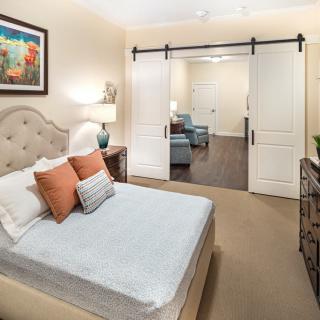 A cozy bedroom with a neatly made bed, featuring colorful pillows and artwork, separated from a sitting area by sliding doors.