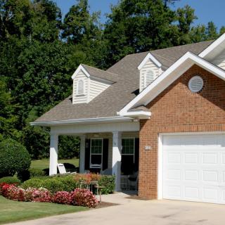 A suburban brick house with a well-maintained lawn and garden, featuring a garage and covered porch.