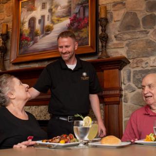 An elderly couple enjoys a meal at a restaurant, while a friendly waiter engages in conversation.