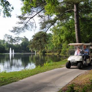 A golf cart driving along a scenic pathway by the lake, surrounded by trees and a fountain.