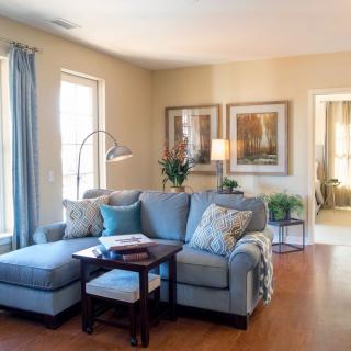 A cozy living room with a blue sectional sofa, wooden flooring, and natural light streaming through large windows.