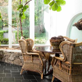 A cozy dining area with wicker chairs and a table, surrounded by large glass windows and indoor plants.