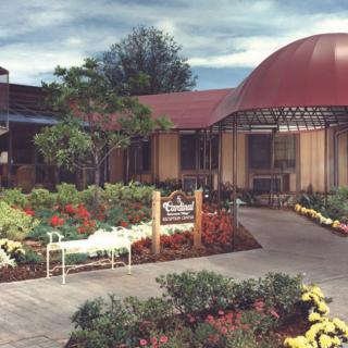 A scenic garden with a variety of plants and flowers in front of a building with a red domed roof, featuring a pathway and bench.