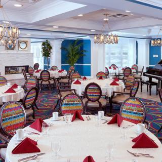 An elegantly decorated dining room with round tables set with red napkins and colorful chairs, featuring a piano in the corner.