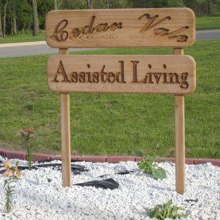 A wooden sign for Cedar Vlg Assisted Living, placed in a landscaped area.