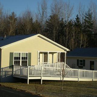 A single-story house with a white accessibility ramp and a front yard, surrounded by trees under clear blue skies.
