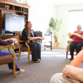 A group of people participating in an indoor exercise session, seated in a circle and holding foam tubes.
