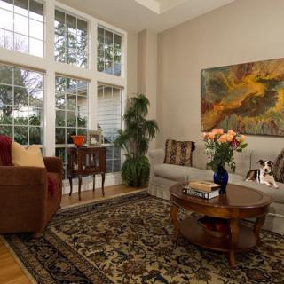 A cozy living room with large windows, a plush sofa, a wooden coffee table adorned with flowers, and vibrant artwork on the wall.