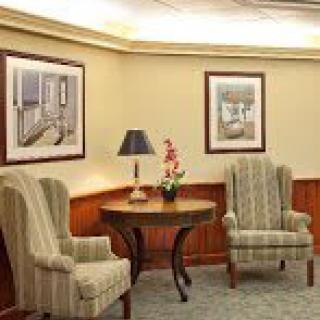 A cozy lobby area with two upholstered chairs, a round table with a lamp and a pink flower, all set against wood-paneled walls with framed pictures.