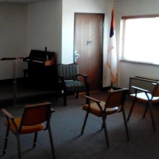 A simple meeting room featuring a few chairs, a piano, and natural light from a window.