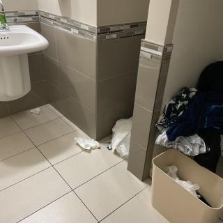 A cluttered bathroom with towels on the floor and a filled trash bin.