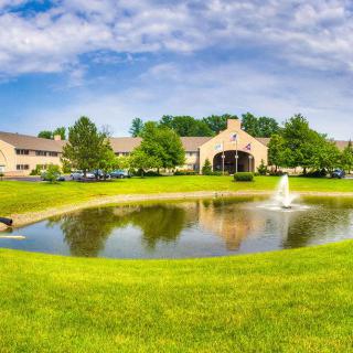A picturesque view of a building surrounded by a pond with a fountain, lush greenery, and a partly cloudy sky.