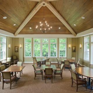 A spacious dining room with several tables and chairs, illuminated by a chandelier and natural light streaming through large windows.
