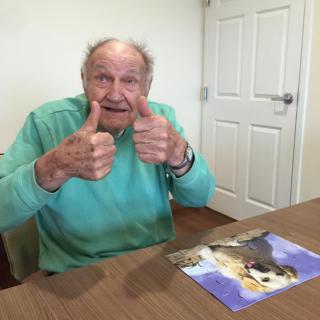 An elderly man giving a thumbs-up while sitting at a table with artwork.
