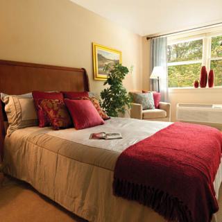 A cozy bedroom with a neatly made bed, adorned with colorful pillows and a burgundy throw, situated next to a window that lets in natural light.