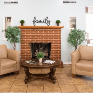 A cozy living room with a brick fireplace, two beige chairs, green indoor plants, and a wooden coffee table.