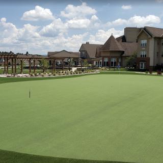 A scenic view of a golf course putting green with a clubhouse in the background under a partly cloudy sky.
