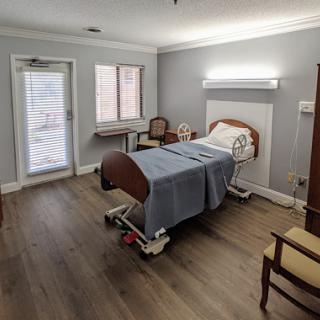 A simple and well-lit hospital patient room featuring a bed, a chair, and windows for natural light.