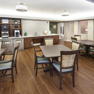 A modern dining room with wooden flooring, featuring multiple tables and chairs, and a kitchen area in the background.