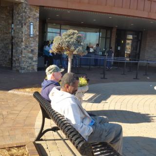 Two people sitting on a bench outside a building on a sunny day.