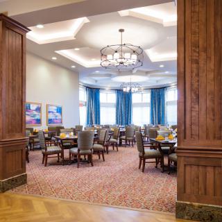 Elegant dining room with neatly arranged tables and chairs, featuring chandeliers and large windows with blue curtains.