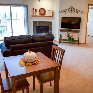 A cozy living room with a dining area features a brown sofa, a wooden table with a decorative pumpkin centerpiece, and a TV set next to a fireplace.