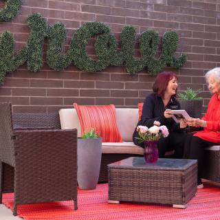 Two women enjoy a conversation in an outdoor seating area with rattan furniture and greenery.