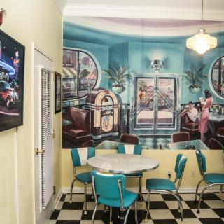 A retro-style diner with a vintage wall mural, checkered floor, and turquoise chairs.