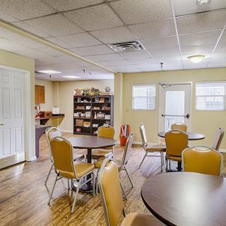 A community room with round tables and chairs set on a wooden floor, featuring windows and a shelving unit in the background.