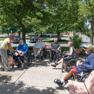 A group of elderly individuals in wheelchairs enjoying a sunny day outdoors, engaging in conversation with a caregiver.