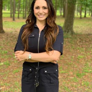 A confident woman posing outdoors with a smile amidst the trees.
