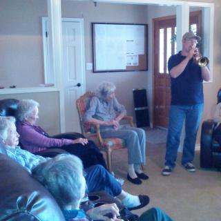 A group of elderly people sitting in a living room, listening to a person playing the trumpet.