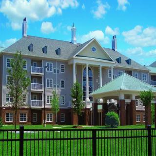 A view of a multi-story residential apartment building with balconies, surrounded by green lawns and bordered by a black fence under a partially cloudy sky.