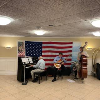 A lively music performance featuring a pianist, guitarist, and bassist in front of an American flag backdrop.