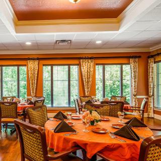 A warmly decorated dining room with round tables set for a meal, featuring large windows with views of greenery.