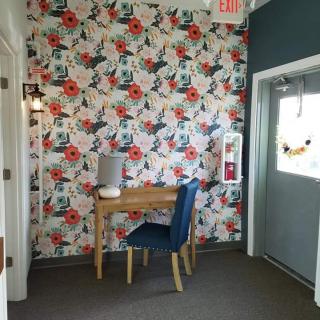 A cozy corner with vibrant floral wallpaper, featuring a small wooden desk and a blue chair, complemented by a stylish lamp.