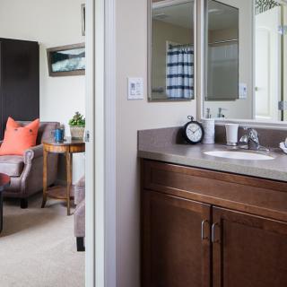 A cozy bedroom with an adjoining bathroom featuring a sink and mirror, along with a comfortable chair and side table.