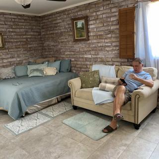 A cozy living room with a man reading on the sofa, adjacent to a bed, featuring brick walls and ample natural light.