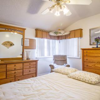 A cozy bedroom featuring wooden furniture, including a dresser with a mirror and a bed. The room is well-lit with natural light from the windows and has a ceiling fan for ventilation.