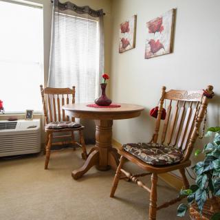A cozy dining nook with a round wooden table surrounded by two matching chairs, adorned with floral wall art and a soft curtain filtering natural light.