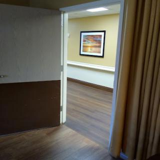 A view through an open doorway into a well-lit, empty room featuring wooden flooring and a piece of wall art.
