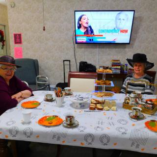 Two people enjoying afternoon tea at a table with snacks and hats, while a television is playing in the background.