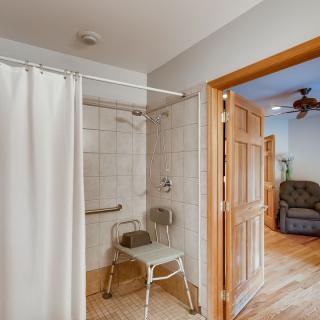 An accessible bathroom with a shower, curtain, and chair, featuring tiled walls and a grab bar near the doorway.