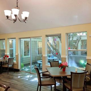 A cozy dining area with tables and chairs, featuring large windows and a chandelier.